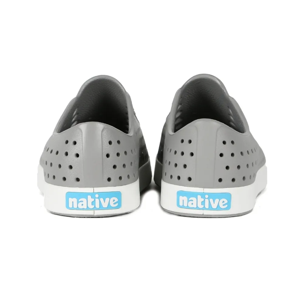 Native รองเท้า Jefferson Pigeon Grey ที่ถ่ายจากด้านหลัง แสดงให้เห็นรองเท้าสีเทาพร้อมโลโก้ Native สีฟ้าบนพื้นรองเท้าสีขาว รองเท้ามีรูระบายอากาศทั่วทั้งคู่ เหมาะสำหรับใส่ในชีวิตประจำวัน